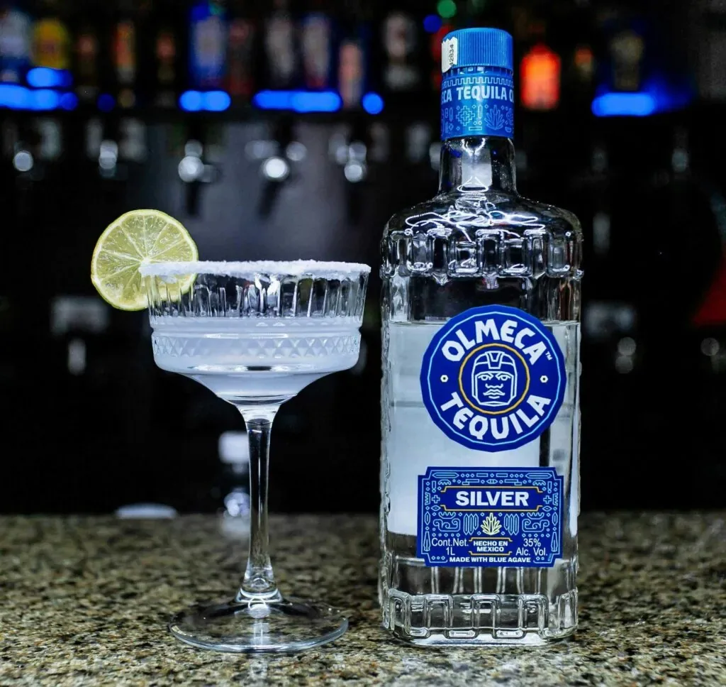A margarita cocktail with a lime slice and Olmeca Tequila bottle on a bar counter.