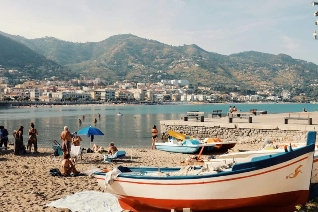 Sunny day at Cefalù beach with rowboats and mountains in view.
