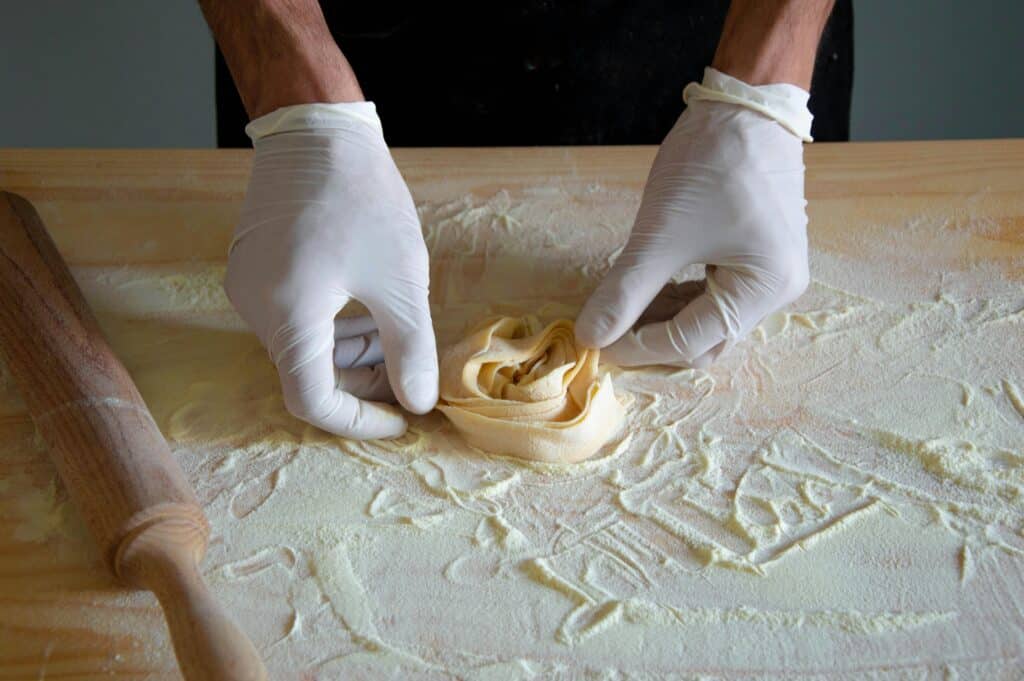 Close-up image of hands wearing gloves making homemade pasta on a wooden surface.