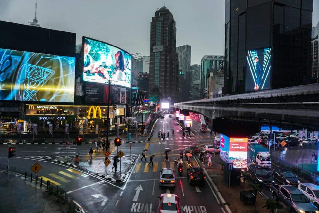 Crowded intersection in Kuala Lumpur at night, with vibrant billboards and traffic.