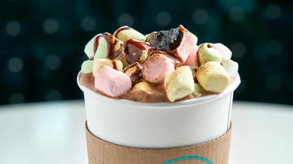 Close-up of hot chocolate topped with colorful marshmallows and chocolate syrup in a to-go cup.