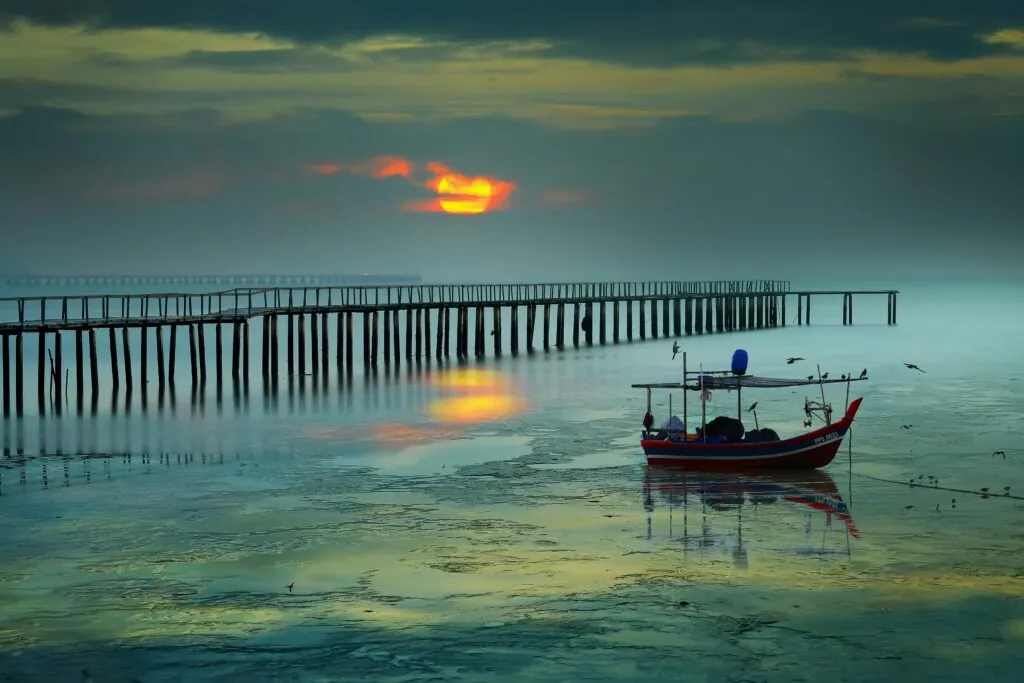 Serene sunrise view with boat and pier in George Town, Malaysia, reflecting vibrant colors on the sea.