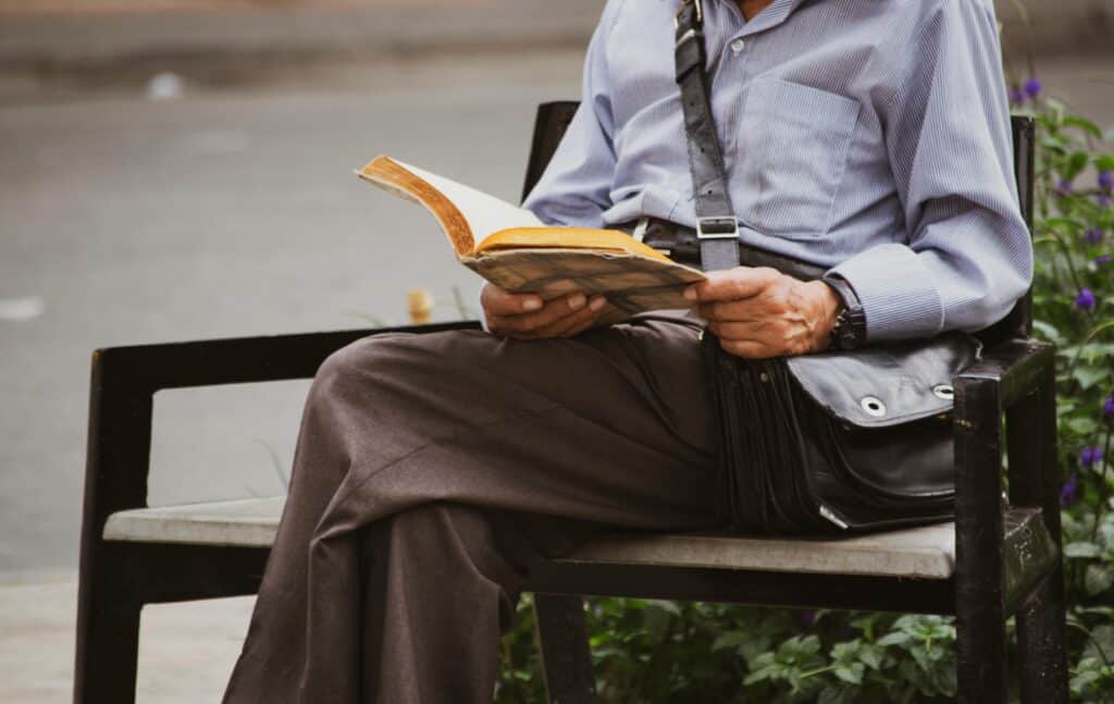 Elderly man enjoying leisure time reading a book on a bench outdoors.