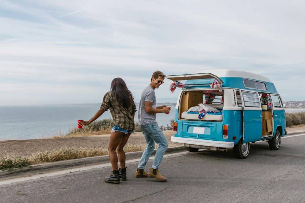 Happy couple dancing by their blue campervan on a scenic coastal road trip.