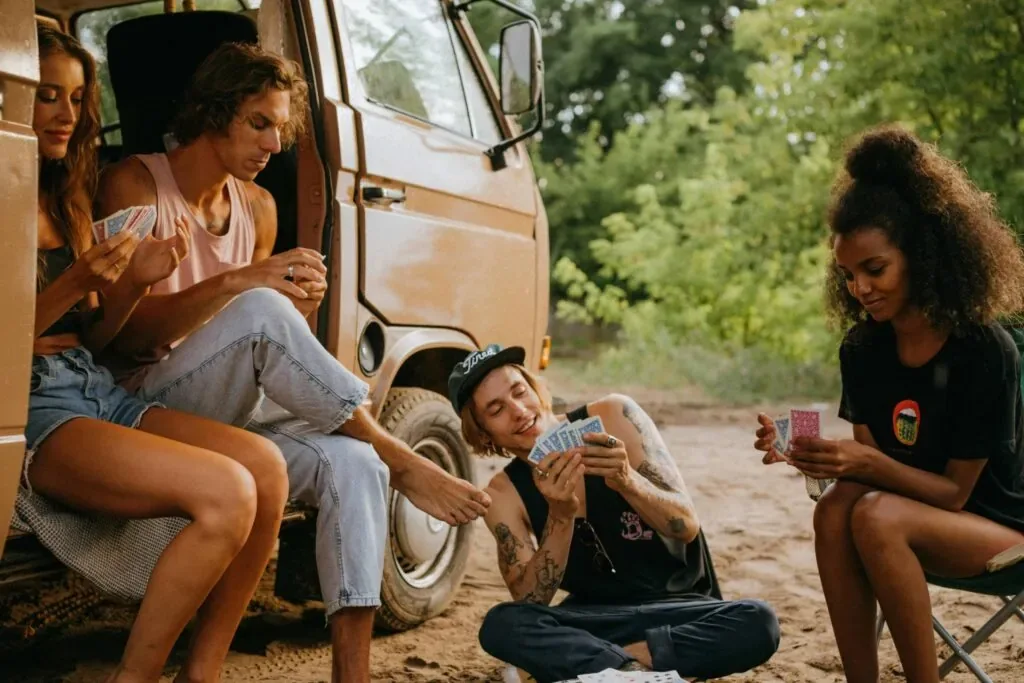 Young campers play cards by a camper van in a forest setting, enjoying a summer adventure.