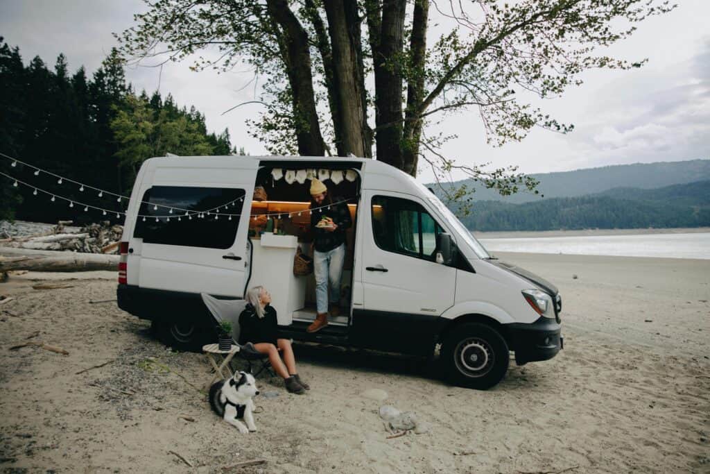 A couple with their dog relax by a camper van on a serene beach, embracing outdoor lifestyle.