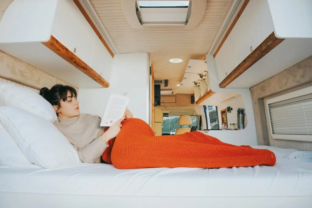 Woman enjoys reading in a cozy camper van bed, wrapped in a warm blanket, capturing the essence of leisure travel.