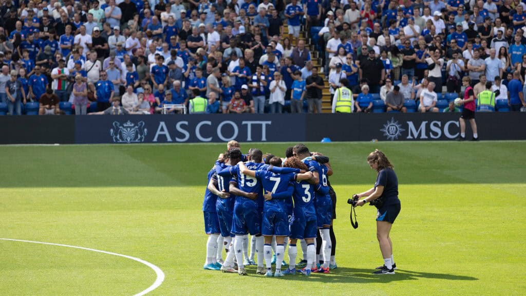 chelsea football club, huddled i a circle on the pitch with a photographer taking photos