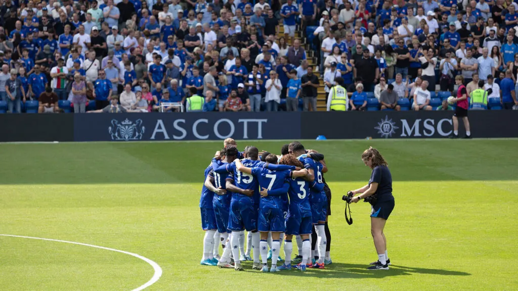 chelsea football club, huddled i a circle on the pitch with a photographer taking photos