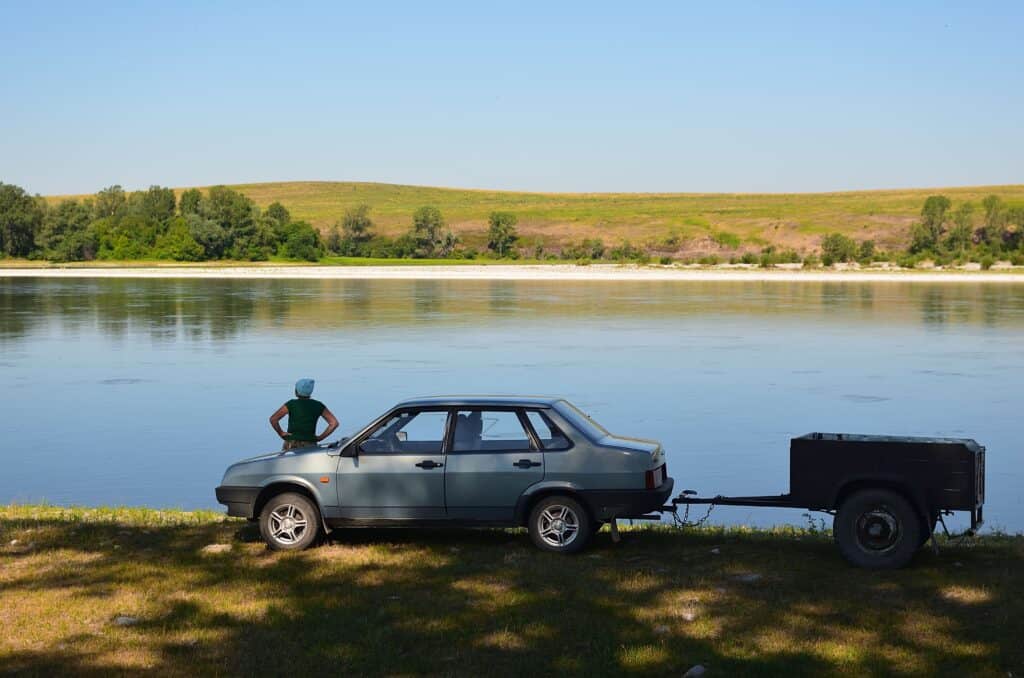 A woman stands beside a car with a trailer near a serene riverbank under a clear sky.