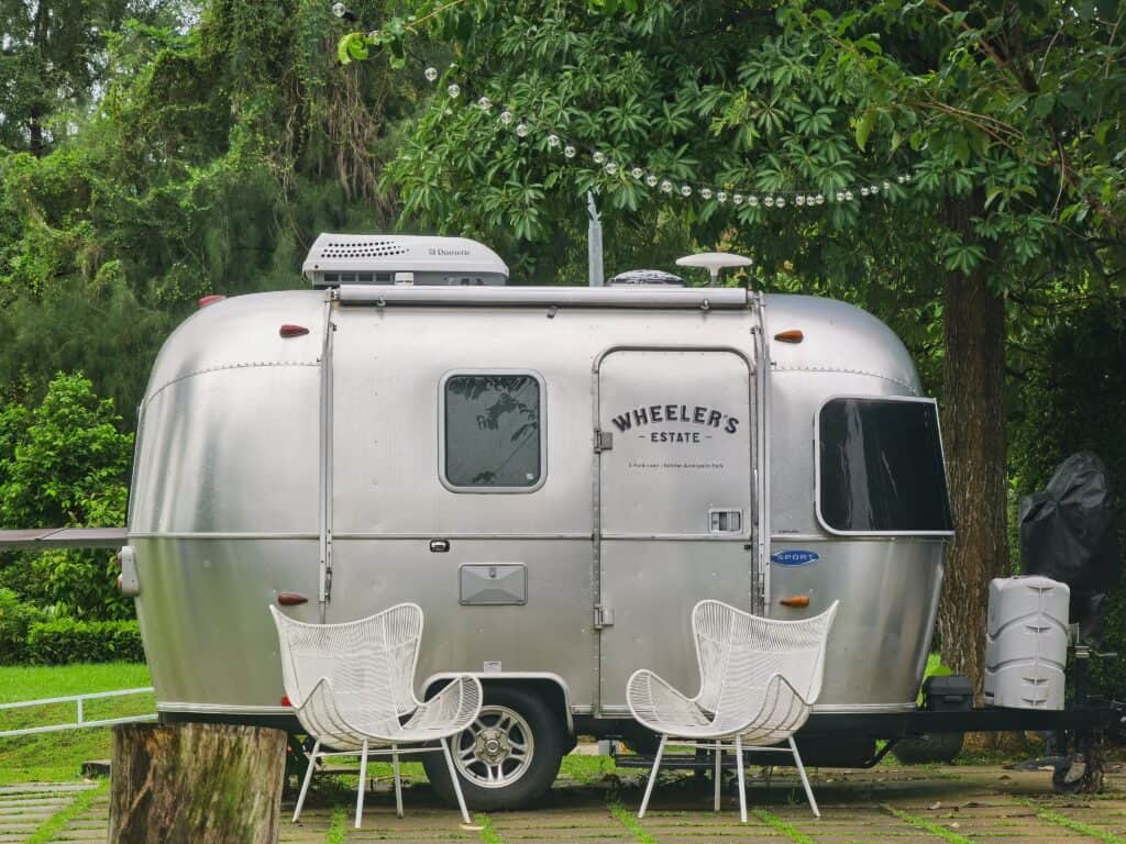 A vintage Airstream caravan parked in a lush Singapore garden with two modern white chairs.