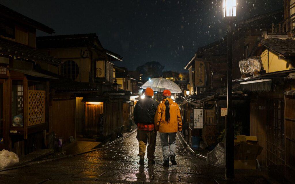 Two people stroll down a rainy street in Kyoto, Japan under an umbrella at night.