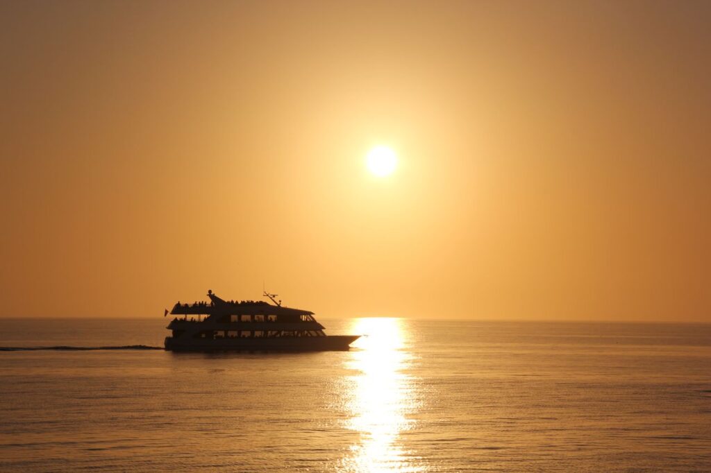 Silhouette of a cruise ship sailing at sunset, reflecting on calm open waters.