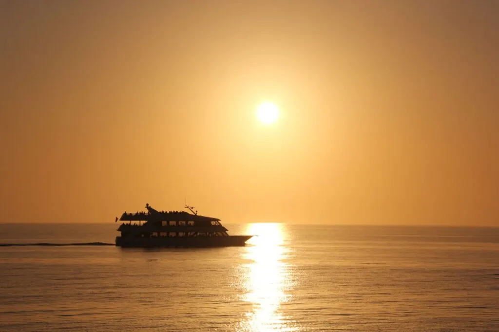 Silhouette of a cruise ship sailing at sunset, reflecting on calm open waters.