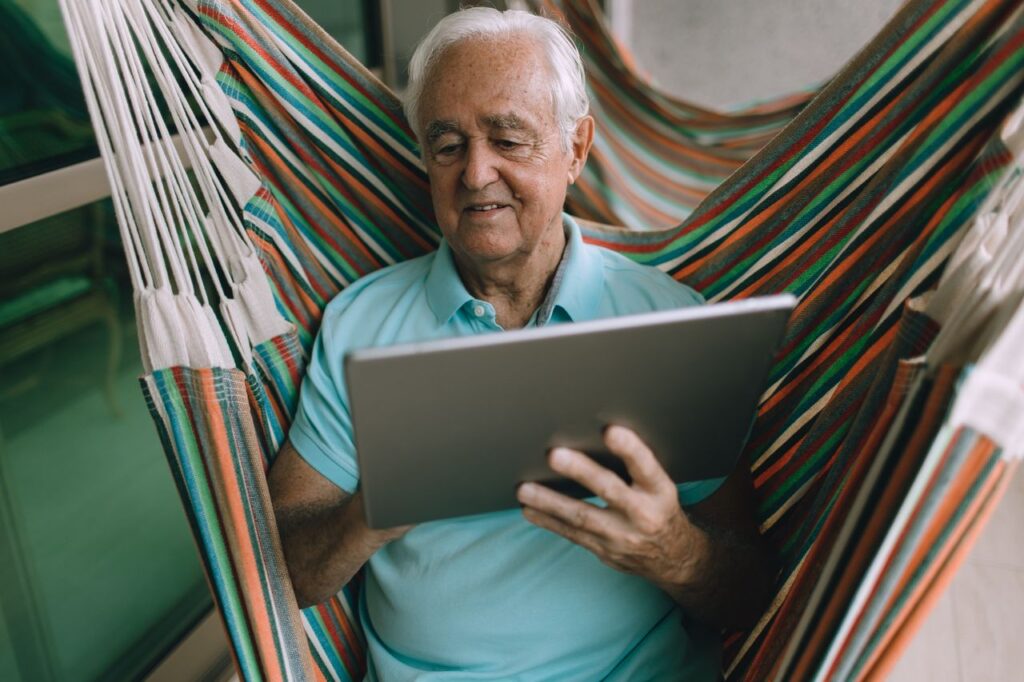 Elderly man enjoying leisure time in a hammock while using a tablet indoors.