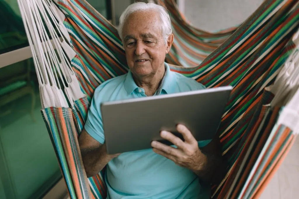 Elderly man enjoying leisure time in a hammock while using a tablet indoors.