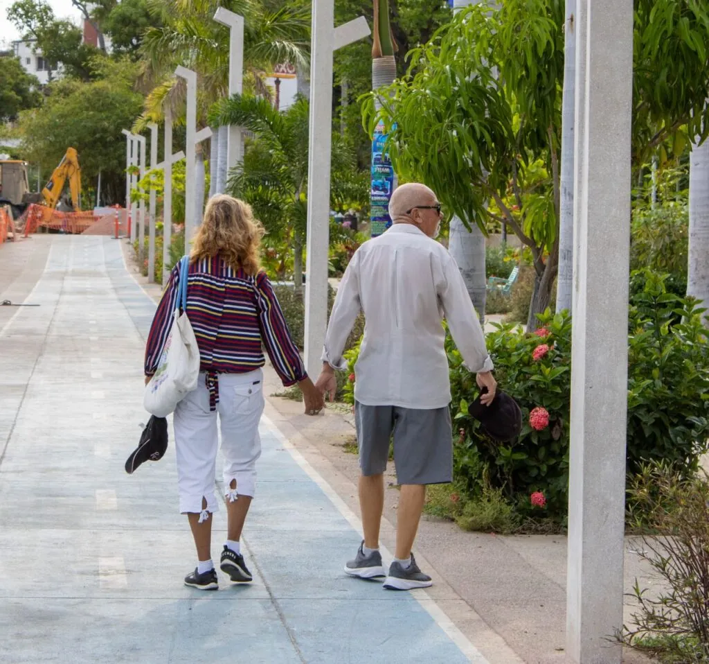 A senior couple walks hand in hand along a garden pathway in Acapulco, Mexico, enjoying a sunny day.