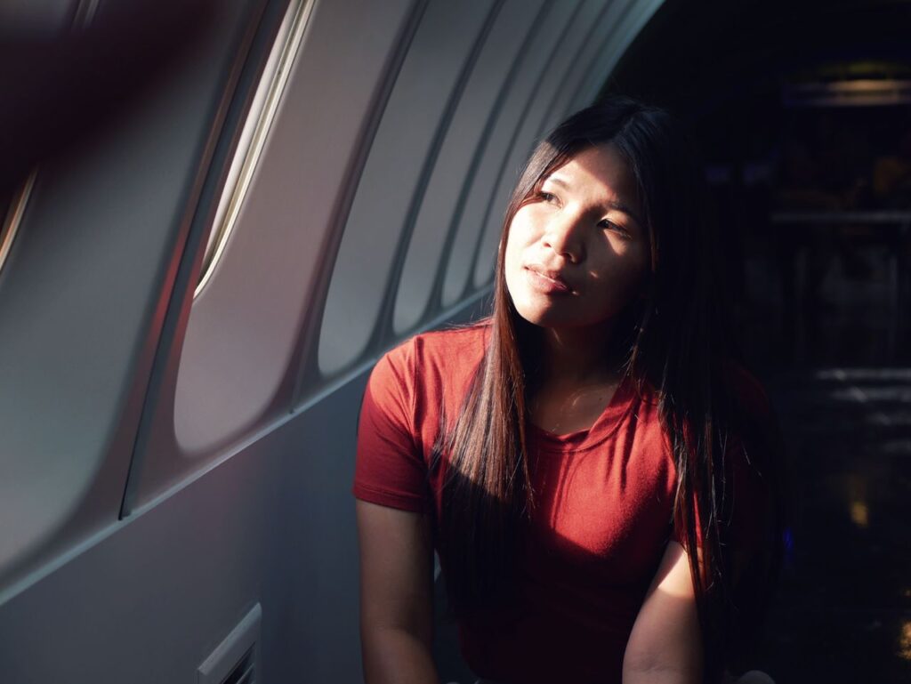 A woman in a red shirt sits in an airplane, gazing thoughtfully out the window as sunlight illuminates her face.
